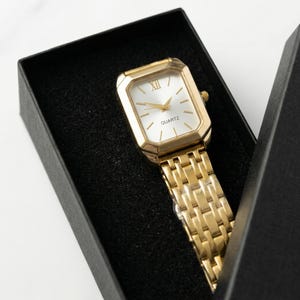 Op de afbeelding: Een goudkleurig quartz horloge met een rechthoekige wijzerplaat en een gouden schakelband, gepresenteerd in een zwarte doos. De wijzerplaat is wit met gouden wijzers en het woord "QUARTZ" eronder.