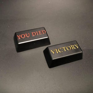 May include: Two black rectangular keycaps with raised lettering. One reads "YOU DIED" in red, the other "VICTORY" in gold. The keycaps are on a dark gray surface.