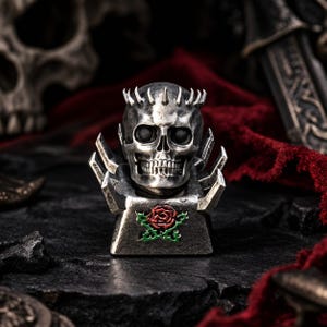 May include: A metallic keycap featuring a detailed human skull with a crown of spikes on top. The base of the keycap is rectangular with sharp, angular protrusions on the sides and a small, colorful red rose with green leaves centered on the front.