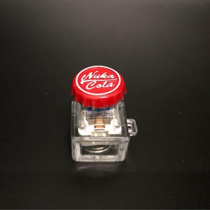 May include: A clear, rectangular, plastic container with a red bottle cap that reads "Nuka Cola" in white lettering. The container has a small handle on the side and contains internal components. The background is black.