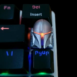 Peut inclure: Un capuchon de clavier en forme de casque de Mandalorien argenté et orange avec une lumière orange qui brille dans la visière. Le capuchon de clavier est sur un clavier noir avec le mot "PgUp" imprimé sur la touche.