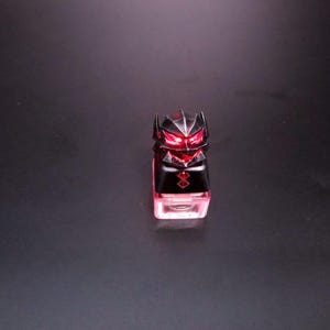 May include: A black and silver keycap with a red glowing design. The keycap has a geometric design with a face-like appearance and a red glowing base. The keycap is likely for a mechanical keyboard.