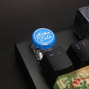 May include: A blue and white Nuka Cola bottle cap keycap on a black keyboard. The keycap is round with the words "Nuka Cola" in white script. Other black keycaps are visible, along with a clear keycap with a koi fish design.