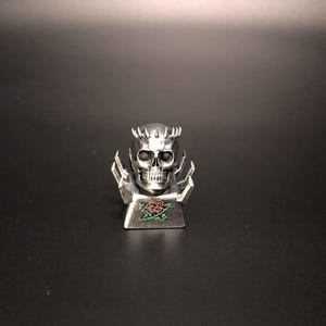 May include: A silver skull-shaped decorative object with a crown and stylized wings. The skull has detailed facial features and is set on a rectangular base with a rose design. The background is a dark gray.