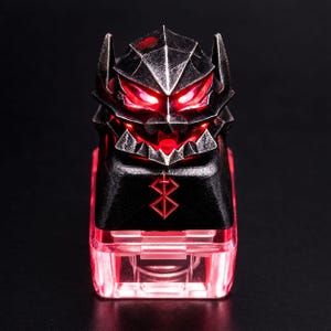 May include: A black mechanical keyboard keycap featuring a sculpted, armoured helmet design with glowing red eyes. The front of the keycap displays a red engraved symbol, and the base is made of a translucent red material that emits a bright light.