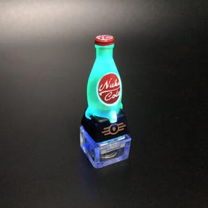 May include: A glowing Nuka Cola bottle figurine with a red cap and a red and white logo. The bottle is teal and sits on a black base with a gold emblem and a clear, blue-lit bottom.