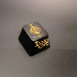 May include: A black keycap featuring gold lettering and a gold emblem. The keycap displays the words "ELDEN RING" in gold. The emblem is a stylized tree with a crown.