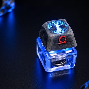 May include: A mechanical keyboard keycap featuring a dark, textured surface with a glowing blue tree of life emblem on top and a red Greek omega symbol on the front face. The base is made of clear, transparent plastic with internal blue lighting.