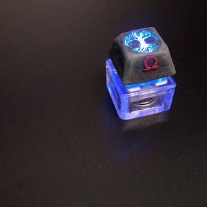 以下が含まれることがあります： 青く光る木の模様と赤いΩ（オメガ）のシンボルが特徴的なカスタムキーキャップ。暗い背景の中で、透明で青く光るベースの上に配置されています。メカニカルキーボード用のユニークなアクセサリーです。