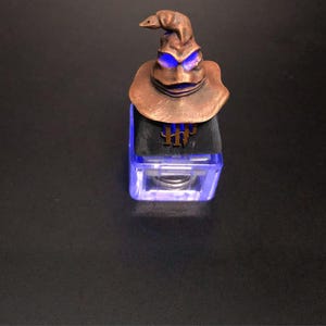 Peut inclure: Une figurine du Choixpeau magique en bronze avec des yeux bleus brillants est posée sur un cube transparent et éclairé. Le cube affiche les lettres "HH" en or. La figurine est une réplique détaillée.