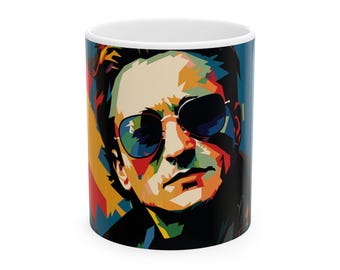 May include: A white ceramic mug with a colorful pop art illustration of a man wearing sunglasses. The man's face is the main focus of the image, with his sunglasses and hair highlighted in bold colors.