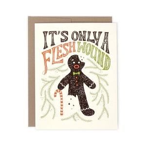 May include: A humorous Christmas card with the text "It's Only a Flesh Wound." The card features a gingerbread man with a bite taken out of him, a candy cane, and green sprigs. The card is on a cream background with a brown envelope.