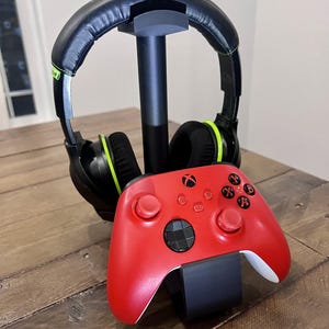 May include: A black and green gaming headset sits on a black stand with a red Xbox controller.
