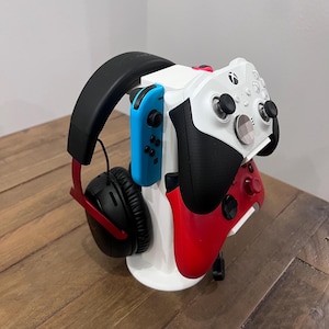 May include: A white stand with a red and black Xbox controller, a blue and black Nintendo Switch controller, and a black headset with red accents.