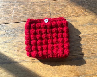May include: A red knitted pot holder with a white button in the centre. The pot holder is made of a textured fabric with a grid pattern.