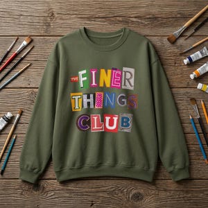 Könnte beinhalten: Ein olivgrünes Sweatshirt mit Rundhalsausschnitt und einem Grafikdesign mit dem Text THE FINER THINGS CLUB. Die Buchstaben sind im Stil eines Erpresserbriefs gestaltet, wobei jeder Buchstabe auf einem bunten, gemusterten Hintergrund liegt.
