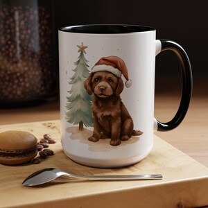 Op de afbeelding: Witte keramische mok met een zwarte handgreep en rand. De mok heeft een aquarel illustratie van een bruine puppy met een kerstmuts, met een kerstboom en een ster. Een lepel en een macaron liggen op een houten oppervlak.