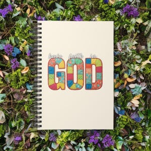 May include: A spiral-bound notebook with a cream-colored cover. The words "have faith in GOD" are printed on the cover in a colorful patchwork design. The notebook is surrounded by greenery and flowers.