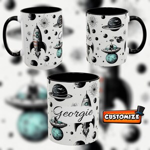 Peut inclure: Mug en céramique blanche avec une anse et un intérieur noirs, orné d'un motif sur le thème de l'espace avec des fusées, des planètes et un OVNI. Le nom "Georgie" est écrit sur le mug. Le mot "Personnaliser" est sur un bouton.