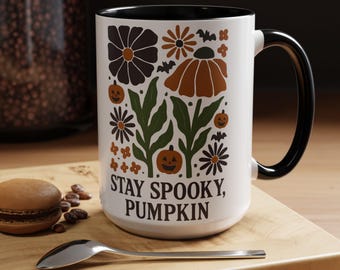 Op de afbeelding: Witte keramische mok met een zwart handvat en rand, versierd met herfstbloemen en pompoen illustraties. De mok heeft de tekst "STAY SPOOKY, PUMPKIN" in een vetgedrukte letter, perfect voor Halloween.