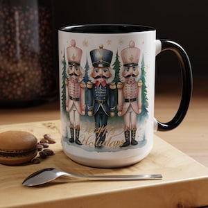 Könnte beinhalten: Weißer Keramikbecher mit schwarzem Griff und Innenraum, mit einer Aquarellillustration von drei Nussknackersoldaten. Der Becher trägt den Text "Happy Holidays" in goldener Schrift und hat ein festliches Thema.