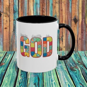 May include: White ceramic mug with a black interior and handle. The mug features the words "have faith in GOD" in a colorful patchwork design. The background is a wooden surface with vertical planks.