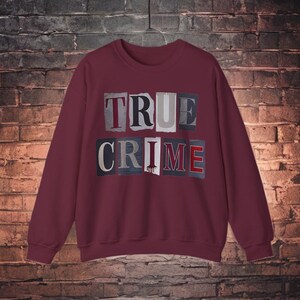 Può includere: Una felpa a maniche lunghe color bordeaux con la scritta TRUE CRIME stampata in stile lettera di riscatto. Le lettere sono visualizzate in singoli blocchi rettangolari con caratteri e colori vari, tra cui rosso, nero, bianco e grigio, sul petto.