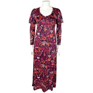 May include: A floor-length dress featuring a dark navy blue base with a vibrant floral pattern in shades of red, orange, pink, and white. The garment includes long sleeves, a V-neckline with a small tie detail, and a cape-like overlay at the shoulders.