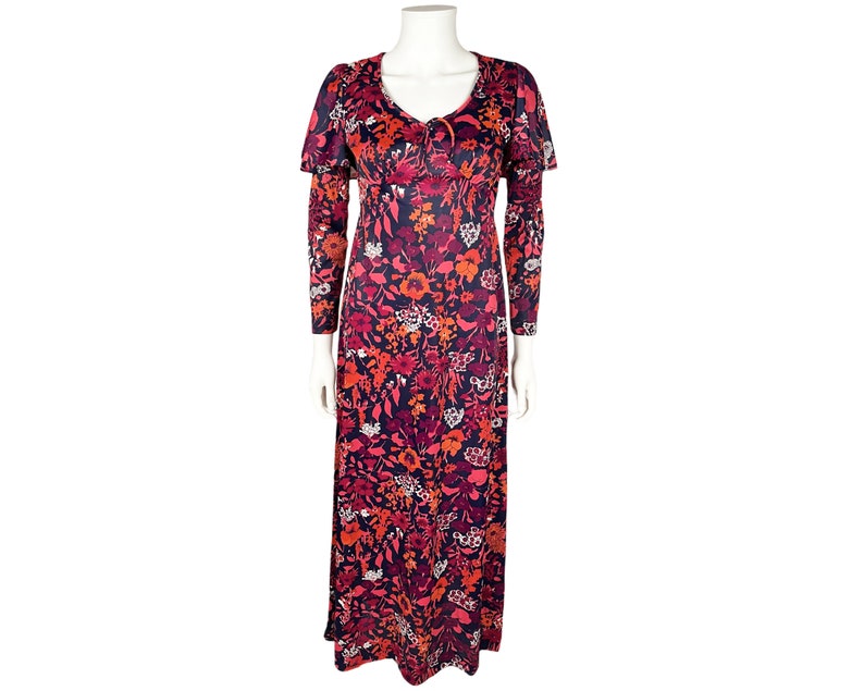 May include: A floor-length dress featuring a dark navy blue base with a vibrant floral pattern in shades of red, orange, pink, and white. The garment includes long sleeves, a V-neckline with a small tie detail, and a cape-like overlay at the shoulders.