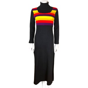 Op de afbeelding: Een lange zwarte jurk met een coltrui en lange mouwen. De borst heeft horizontale strepen in rood, geel en paars. De jurk heeft aan elke kant een split en wordt op een mannequin gepresenteerd.