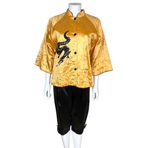 May include: A gold satin jacket with a black dragon design and black buttons. The jacket has a quilted pattern and a mandarin collar. The outfit includes black satin cropped pants. The jacket's sleeves are three-quarter length.