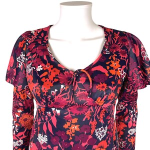 May include: A long-sleeved dress featuring a dark navy blue base with a vibrant floral pattern in shades of red, pink, and orange. The garment includes a V-neckline with a central tie detail and ruffled flutter sleeves that extend over the shoulders.