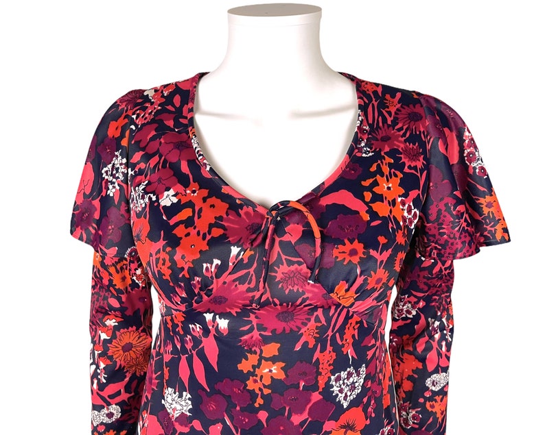May include: A long-sleeved dress featuring a dark navy blue base with a vibrant floral pattern in shades of red, pink, and orange. The garment includes a V-neckline with a central tie detail and ruffled flutter sleeves that extend over the shoulders.