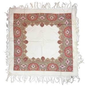 May include: A square off-white shawl featuring a decorative border with intricate paisley patterns in shades of red, gold, and olive green. The fabric is finished with long, knotted white fringe along all four edges of the garment.