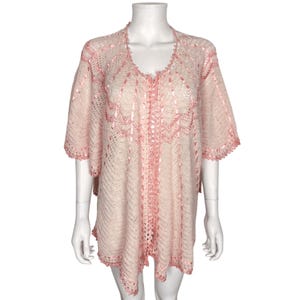 May include: A light pink crochet cardigan featuring an open-knit pattern with darker pink ribbon detailing woven throughout the fabric. The garment has short sleeves, a scalloped edge along the neckline and hem, and a loose, flowing silhouette.