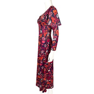 May include: A floor-length dress featuring a dark navy blue base with a vibrant floral pattern in shades of red, orange, pink, and white. The garment has long sleeves, a V-neckline, and a cape-like overlay detail on the shoulders and upper back.