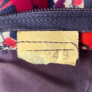 May include: A fabric garment featuring a dark blue zipper and a floral pattern in shades of red, pink, and blue. A rectangular, light-colored paper tag is stitched to the fabric near the zipper, displaying the handwritten number 549 in pencil.