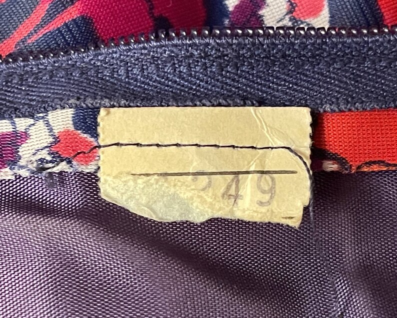 May include: A fabric garment featuring a dark blue zipper and a floral pattern in shades of red, pink, and blue. A rectangular, light-colored paper tag is stitched to the fabric near the zipper, displaying the handwritten number 549 in pencil.