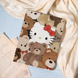 May include: A rectangular tablet case with a repeating pattern of teddy bears in shades of brown and tan. A large image of Hello Kitty with a red bow is centered on the case. The background is a cream-coloured fabric.
