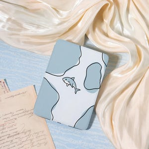 May include: A light blue and white case with a shark illustration. The case has a pattern of light blue shapes outlined in black. The shark is drawn in a simple, cartoon style. The case is likely for a tablet or e-reader.