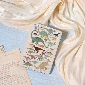 May include: A rectangular, off-white case featuring a colorful illustration of various dinosaurs. The dinosaurs are depicted in shades of green, brown, yellow, and purple. The case is set against a backdrop of cream-colored fabric and vintage paper.