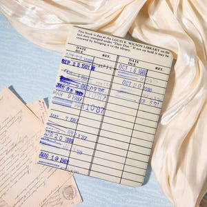 May include: A vintage library card with blue ink stamps and handwritten dates, including "Date Due" and "Ret." The card is on a light blue surface with cream-colored fabric in the background. Old letters are also visible.