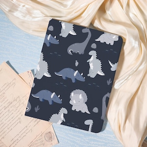 May include: A dark blue tablet case featuring a pattern of cartoon dinosaurs in shades of gray and blue. The dinosaurs include Triceratops, Stegosaurus, and Brontosaurus. The case is rectangular and has rounded corners.