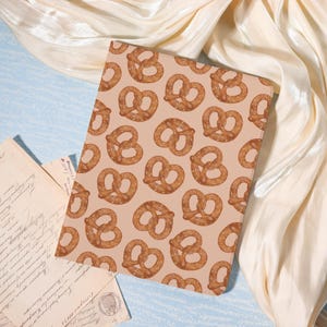 May include: A notebook with a repeating pattern of brown pretzels on a tan background. The notebook is rectangular and lies on a light blue surface with cream-colored fabric and vintage letters in the background.