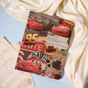 May include: A rectangular print featuring Lightning McQueen from the film Cars, the number 95, and the text "CARS 2006." The print has a collage design with various images and text elements, set against a cream-coloured fabric.
