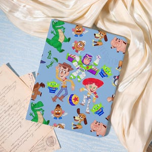 May include: A light blue tablet case featuring cartoon characters from the Toy Story franchise. The case displays images of Woody, Buzz Lightyear, Jessie, Rex, Slinky Dog, Hamm, and the Little Green Men. The design is a colorful illustration.