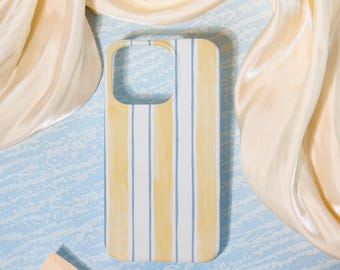 May include: A phone case with a vertical striped pattern in yellow, white, and light blue. The case is rectangular with rounded corners and a cutout for the camera. The background includes a light blue surface and a flowing, cream-colored fabric.
