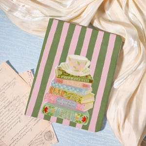 May include: A notebook with a green and pink striped cover featuring a stack of books and a teacup illustration. The books have floral designs and titles like "Wild Flowers" and "Anne of Green Gables". The teacup is pink with a floral pattern.