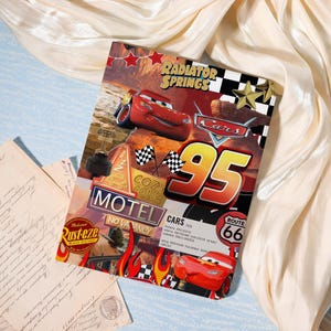 May include: A colorful Cars-themed notebook featuring Lightning McQueen and the number 95. The design includes the words "Radiator Springs", "Cars", "Motel", and "Route 66". The notebook is decorated with checkered flags and flames.