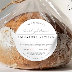 Op de afbeelding: Een rond brood van zuurdesem in een doorzichtige plastic zak, vastgebonden met touw. Het brood heeft een goudbruine korst en een witte, bloemige bovenkant. Een wit etiket op de zak luidt "Sourdough Bread, Signature Artisan". De tekst "Works perfectly for bread bags" is ook zichtbaar.